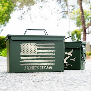 May include: Two olive green metal storage boxes. The larger box features a distressed American flag design and the name "JAMES RYAN" in silver. The smaller box has a design of flying birds. Both have handles.