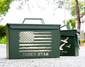 May include: Two olive green metal storage boxes. The larger box features a distressed American flag design and the name "JAMES RYAN" in silver. The smaller box has a design of flying birds. Both have handles.