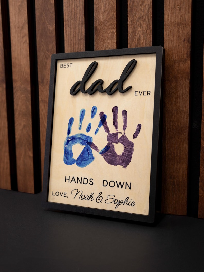 May include: A framed wooden sign with the words "BEST dad EVER" in black 3D lettering. Below are two handprints, one blue and one purple, with the text "HANDS DOWN LOVE, Noah & Sophie".