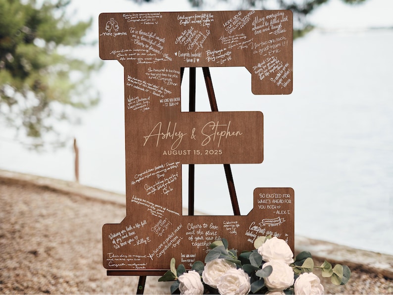 K&ouml;nnte beinhalten: Ein gro&szlig;es Hochzeitszeichen aus Holz in Form des Buchstabens "E" mit Unterschriften, den Namen "Ashley & Stephen" und dem Datum "15. August 2025". Das Schild steht auf einer Holzstaffelei und ist mit wei&szlig;en Blumen geschm&uuml;ckt.