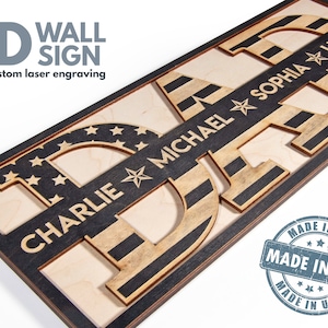 May include: A 3D wooden wall sign spelling "DAD" with an American flag design. The letters are layered and each has a name engraved on it: Charlie, Michael, Sophia, and Lindsay. The sign is framed in black wood and has a "Made in USA" stamp.