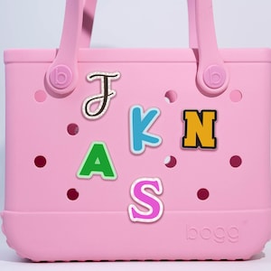 May include: A light pink tote bag with a handle and the word "bogg" printed on the bottom. The bag has several colorful letter charms spelling out the name "J.A.K.N.S.". The letters are in various colors and styles.