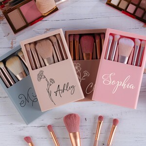 May include: Makeup brush sets in pastel-colored boxes, each containing multiple brushes. The boxes are personalized with names in cursive script. The image also shows eyeshadow palettes and individual makeup brushes.