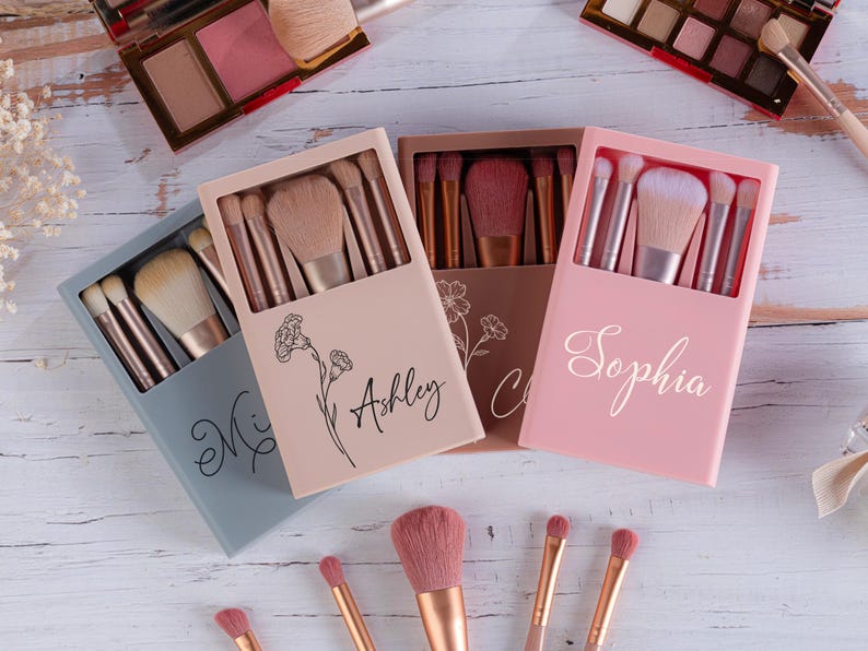 May include: Makeup brush sets in pastel-colored boxes, each containing multiple brushes. The boxes are personalized with names in cursive script. The image also shows eyeshadow palettes and individual makeup brushes.