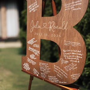 K&ouml;nnte beinhalten: Ein gro&szlig;er Holzbuchstabe "B" mit den Namen "Julia & Ronald" und dem Datum "15. Juli 2025" eingraviert. Das Holzschild steht auf einer Holzstaffelei und ist mit handschriftlichen Notizen bedeckt.