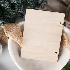 May include: A blank wooden sign with metal rings for hanging, surrounded by hessian ribbon and a white bowl. The sign is sitting on a white surface with a green pine branch and a gold bauble in the background.