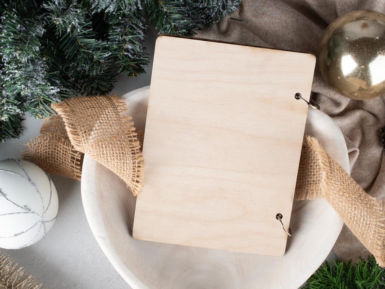 May include: A blank wooden sign with metal rings for hanging, surrounded by hessian ribbon and a white bowl. The sign is sitting on a white surface with a green pine branch and a gold bauble in the background.