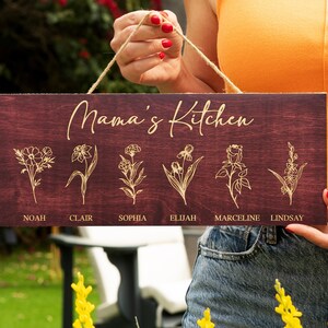 Puede incluir: Un letrero de madera con el texto "Mama's Kitchen" en letras doradas. El letrero tiene seis dise&ntilde;os florales con nombres debajo de cada dise&ntilde;o: Noah, Clair, Sophia, Elijah, Marceline y Lindsay.