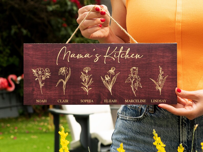 Puede incluir: Un letrero de madera con el texto "Mama's Kitchen" en letras doradas. El letrero tiene seis dise&ntilde;os florales con nombres debajo de cada dise&ntilde;o: Noah, Clair, Sophia, Elijah, Marceline y Lindsay.
