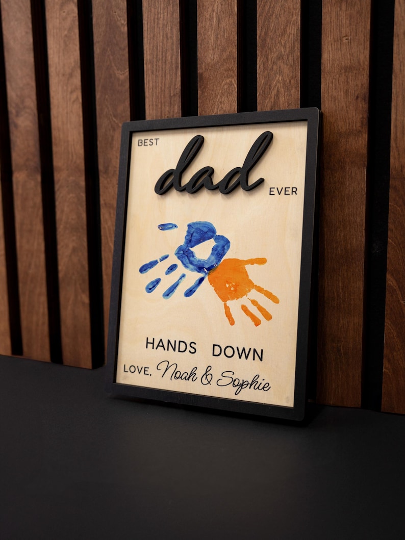May include: A framed wooden sign with the words "Best Dad Ever" in black 3D lettering. The sign features blue and orange handprints and the text "Hands Down Love, Noah & Sophie."