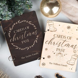 May include: Two wooden photo albums with a rustic brown and natural wood finish. The albums are decorated with a floral wreath and the text "Cards of Christmas Past". The albums are labelled with the names "The Taylor Family" and "The William Family".