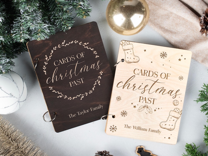 May include: Two wooden photo albums with a rustic brown and natural wood finish. The albums are decorated with a floral wreath and the text "Cards of Christmas Past". The albums are labelled with the names "The Taylor Family" and "The William Family".