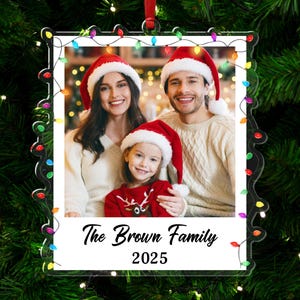 Pode incluir: Um enfeite de Natal de acr&iacute;lico transparente com uma foto de fam&iacute;lia usando gorros de Papai Noel. O enfeite tem uma borda decorativa com luzes coloridas e o texto "The Brown Family 2025". Ele pende de uma fita vermelha.