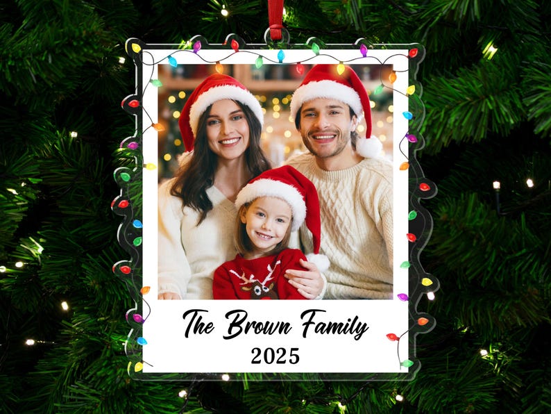 Pode incluir: Um enfeite de Natal de acr&iacute;lico transparente com uma foto de fam&iacute;lia usando gorros de Papai Noel. O enfeite tem uma borda decorativa com luzes coloridas e o texto "The Brown Family 2025". Ele pende de uma fita vermelha.