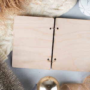 May include: A blank, wooden, two-page journal with metal rings. The journal is open and lying flat on a light grey surface. There is a white knitted sweater, a gold tassel, and a white decorative ball in the background.