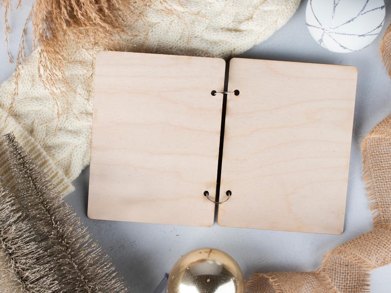 May include: A blank, wooden, two-page journal with metal rings. The journal is open and lying flat on a light grey surface. There is a white knitted sweater, a gold tassel, and a white decorative ball in the background.