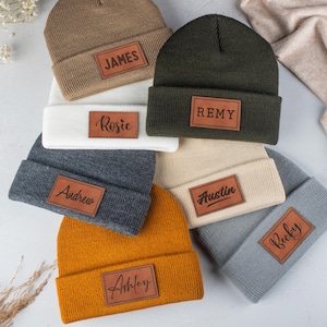 May include: A collection of seven knit beanies in various colors including tan, olive green, white, dark gray, cream, mustard yellow, and light gray. Each beanie features a rectangular brown leather patch with a custom name engraved in black.