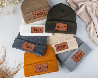 Puede incluir: Una colección de siete gorros de punto en varios colores, incluyendo tostado, verde oliva, blanco, gris oscuro, crema, amarillo mostaza y gris claro. Cada gorro presenta un parche rectangular de cuero marrón con un nombre grabado en negro.