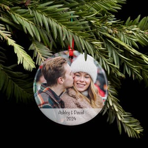 May include: A round, clear glass ornament with a photo of a couple kissing. The ornament has a white background with the text "Ashley & David 2024" printed on it.