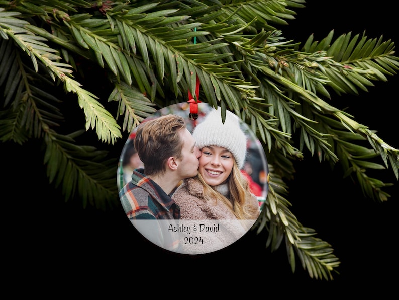 May include: A round, clear glass ornament with a photo of a couple kissing. The ornament has a white background with the text "Ashley & David 2024" printed on it.
