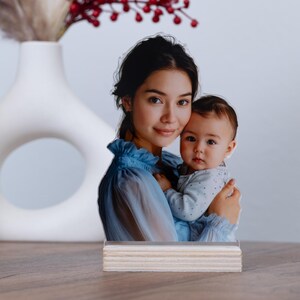 May include: A photo stand featuring a mother and baby. The image is a cutout of a woman in a blue dress holding a baby. The stand is made of light-colored wood and sits on a wooden surface.
