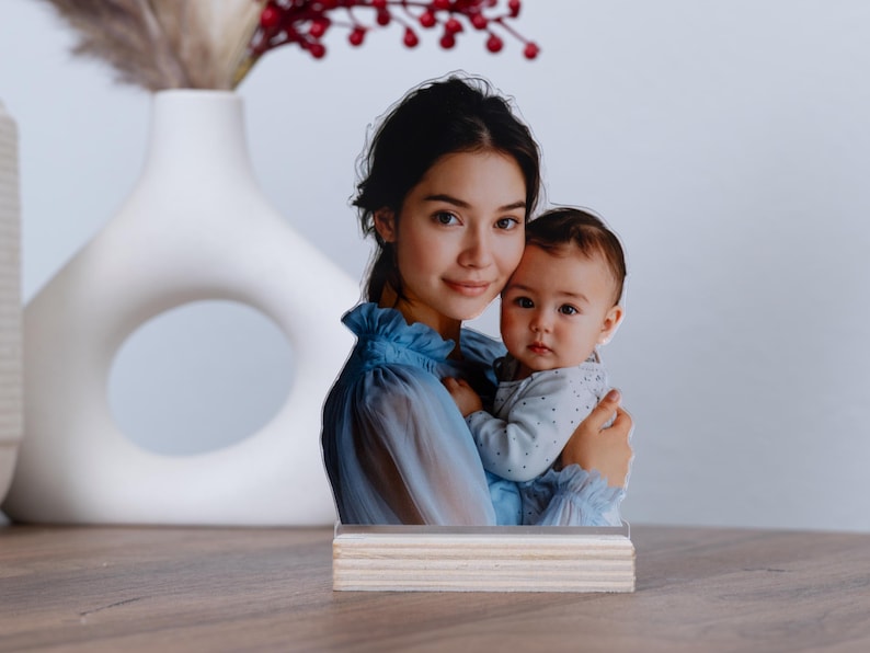 May include: A photo stand featuring a mother and baby. The image is a cutout of a woman in a blue dress holding a baby. The stand is made of light-colored wood and sits on a wooden surface.