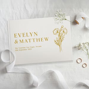 Puede incluir: Un libro de visitas de boda blanco con letras doradas que dicen "EVELYN & MATTHEW". El libro tambi&eacute;n incluye el lugar y la fecha de la boda. Un dise&ntilde;o floral dorado est&aacute; en el lado derecho. Cinta blanca y flores en la imagen.