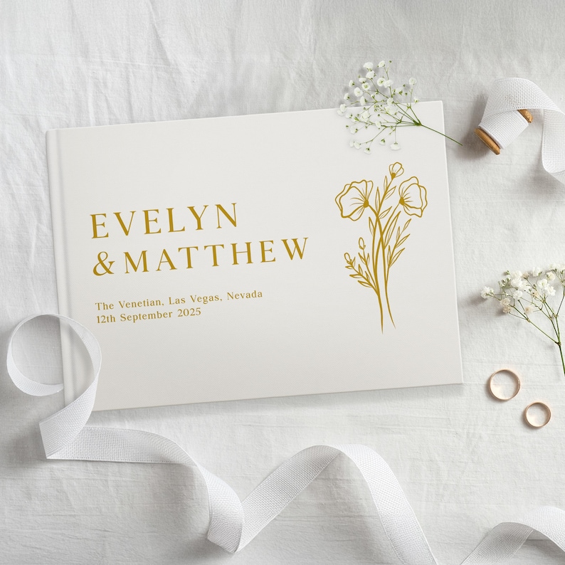 Puede incluir: Un libro de visitas de boda blanco con letras doradas que dicen "EVELYN & MATTHEW". El libro tambi&eacute;n incluye el lugar y la fecha de la boda. Un dise&ntilde;o floral dorado est&aacute; en el lado derecho. Cinta blanca y flores en la imagen.