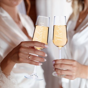 Puede incluir: Dos copas de champ&aacute;n llenas de un l&iacute;quido dorado, levantadas en un brindis de celebraci&oacute;n. Las copas est&aacute;n personalizadas con nombres y fechas. Una dice "Madison Bride 05.08.2022" y la otra "Theresa Maid of Honor 05.08.2022".