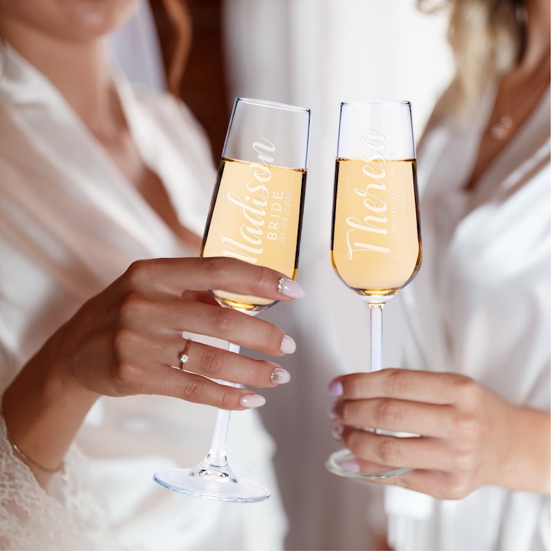 Puede incluir: Dos copas de champ&aacute;n llenas de un l&iacute;quido dorado, levantadas en un brindis de celebraci&oacute;n. Las copas est&aacute;n personalizadas con nombres y fechas. Una dice "Madison Bride 05.08.2022" y la otra "Theresa Maid of Honor 05.08.2022".