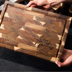 May include: A rectangular wooden chopping board with a juice groove and end-grain butcher block design. The board is engraved with "The Martins" and the date 01.31.2021 in a script font. The board is held by two hands.