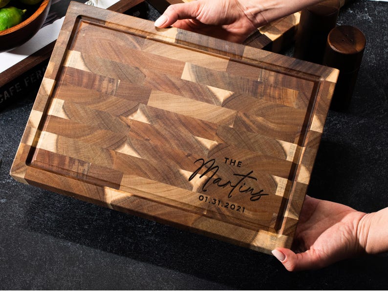 May include: A rectangular wooden chopping board with a juice groove and end-grain butcher block design. The board is engraved with "The Martins" and the date 01.31.2021 in a script font. The board is held by two hands.