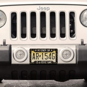 Puede incluir: Un Jeep blanco con una placa de matr&iacute;cula de camuflaje que dice "MY OTHER CAR IS A SIEGE TANK" y el n&uacute;mero de placa "AR 154G".