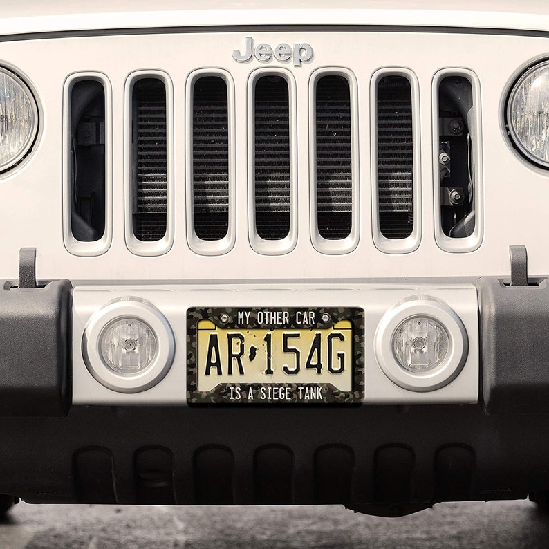 Puede incluir: Un Jeep blanco con una placa de matr&iacute;cula de camuflaje que dice "MY OTHER CAR IS A SIEGE TANK" y el n&uacute;mero de placa "AR 154G".