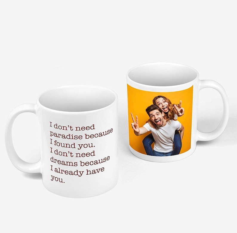 Puede incluir: Taza de cer&aacute;mica blanca con una pareja en la parte posterior, el hombre est&aacute; haciendo un signo de paz y la mujer est&aacute; haciendo un signo de paz con su brazo alrededor del hombre. La parte delantera de la taza tiene el texto "No necesito el para&iacute;so porque te encontr&eacute;. No necesito sue&ntilde;os porque ya te tengo."