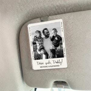 May include: A black and white photo sun visor card with a family portrait. The image features a man holding two children. Text on the card reads "Drive safe, Daddy!" and the names "MICHAEL & RAYMOND".