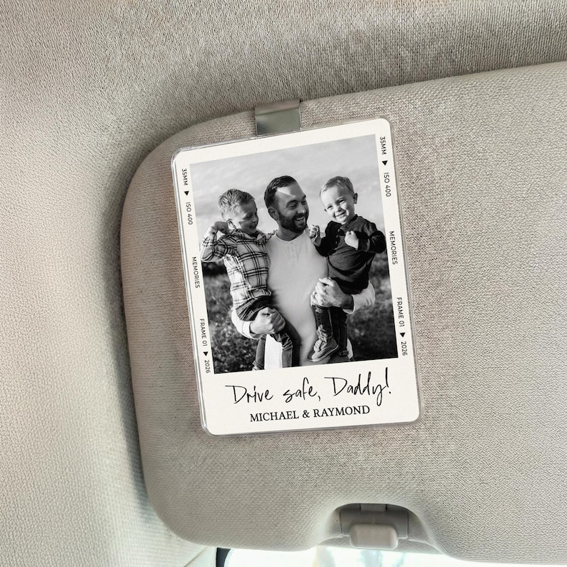 May include: A black and white photo sun visor card with a family portrait. The image features a man holding two children. Text on the card reads "Drive safe, Daddy!" and the names "MICHAEL & RAYMOND".
