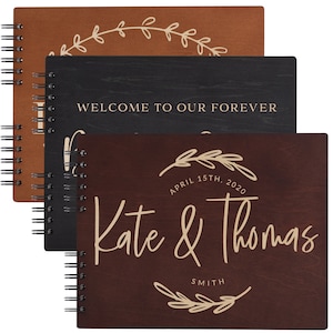 May include: Three wooden guest books with spiral binding. The top book is brown with a leafy wreath design. The middle book is black with the text "Welcome to our forever". The bottom book is brown with the text "Kate & Thomas" and the date "April 15th, 2020".