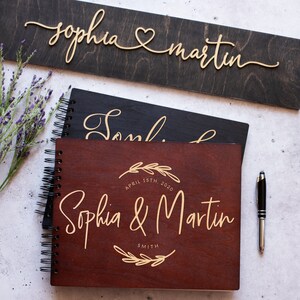 May include: Two wooden guest books with gold lettering. The top book is black with the names "Sophia & Martin" in cursive. The bottom book is brown with the names "Sophia & Martin" in cursive and the date "April 15th, 2020" and the last name "Smith" in a circle with a floral design.