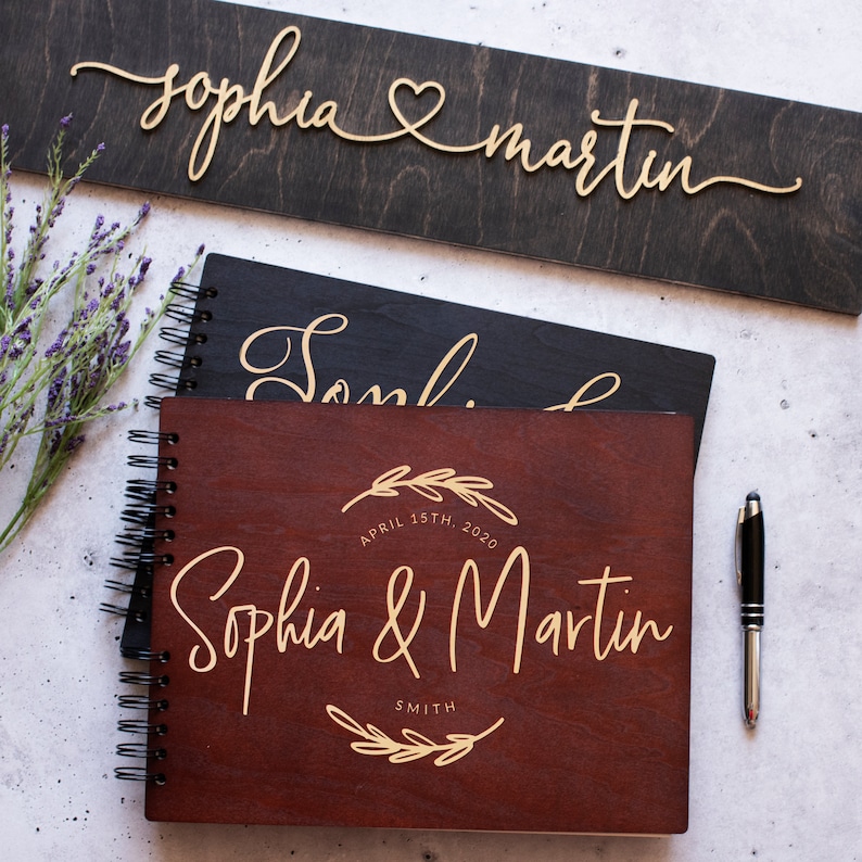 May include: Two wooden guest books with gold lettering. The top book is black with the names "Sophia & Martin" in cursive. The bottom book is brown with the names "Sophia & Martin" in cursive and the date "April 15th, 2020" and the last name "Smith" in a circle with a floral design.