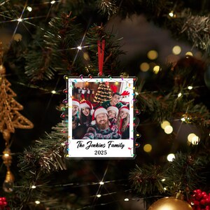 Pode incluir: Enfeite de Natal em acr&iacute;lico transparente com uma foto de fam&iacute;lia no interior, rodeado por uma s&eacute;rie de luzes coloridas. A foto mostra um grupo de pessoas com gorros de Pai Natal. O texto no enfeite diz "The Jenkins Family 2025".