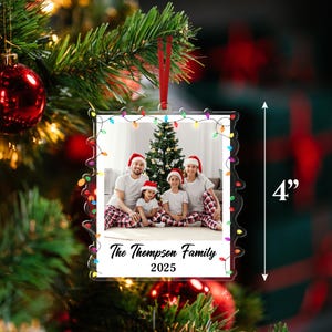 Pode incluir: Um enfeite de Natal de cerca de 10 cm com uma foto de fam&iacute;lia, emoldurado com luzes coloridas. O texto "The Thompson Family 2025" &eacute; vis&iacute;vel. O enfeite est&aacute; pendurado por uma fita vermelha.
