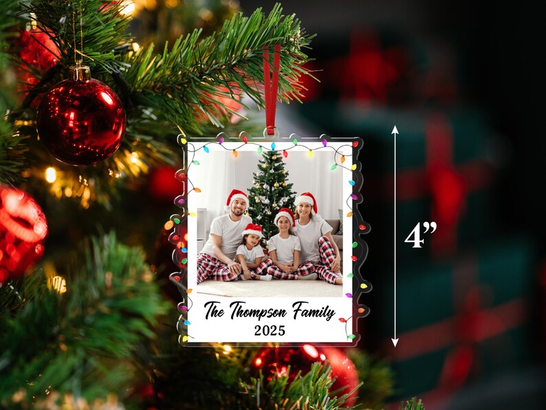 Pode incluir: Um enfeite de Natal de cerca de 10 cm com uma foto de fam&iacute;lia, emoldurado com luzes coloridas. O texto "The Thompson Family 2025" &eacute; vis&iacute;vel. O enfeite est&aacute; pendurado por uma fita vermelha.
