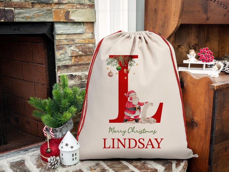 May include: A personalized Christmas gift bag with a red and white Santa Claus illustration and the letter "L" decorated with green holly and gold ornaments. The bag is made of natural canvas fabric and has a red drawstring. The text "Merry Christmas Lindsay" is printed on the bag.