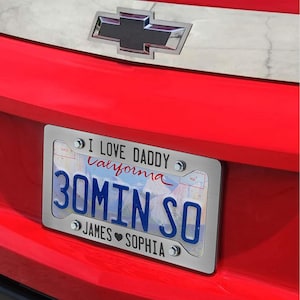 Puede incluir: Una placa de matr&iacute;cula personalizada de plata con el texto "I Love Daddy California 30min So James Sophia" y una imagen de fondo azul y blanco. La placa est&aacute; unida a un coche rojo.