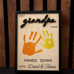May include: A framed wooden sign with the words "BEST grandpa EVER" in black script. The sign features orange and yellow handprints and the text "HANDS DOWN LOVE, David & Fiona."