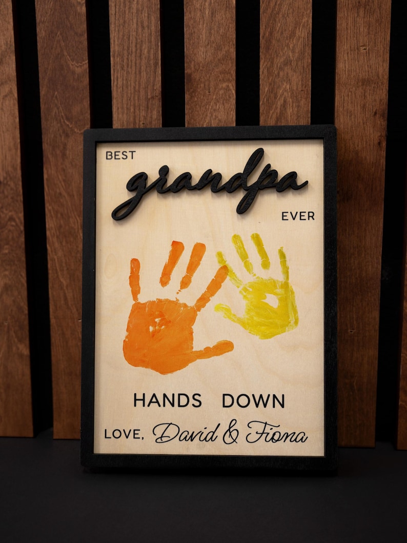 May include: A framed wooden sign with the words "BEST grandpa EVER" in black script. The sign features orange and yellow handprints and the text "HANDS DOWN LOVE, David & Fiona."