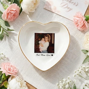 May include: A white heart-shaped ceramic trinket dish with a gold-toned rim. The center features a printed photograph of a person holding a baby, with the black text "Best Mom Ever!" and a small black heart symbol printed below the image.