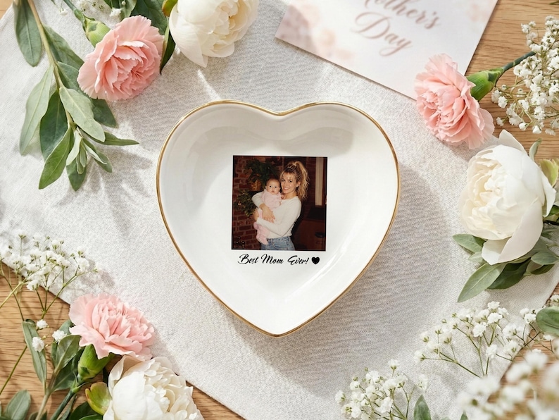 May include: A white heart-shaped ceramic trinket dish with a gold-toned rim. The center features a printed photograph of a person holding a baby, with the black text "Best Mom Ever!" and a small black heart symbol printed below the image.