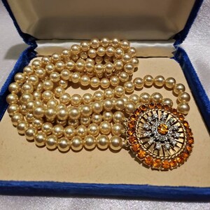 May include: A multi-strand pearl necklace with a large, round, gold-toned pendant featuring a sunburst design with orange and clear rhinestones. The necklace is in a blue velvet box with the words "Originals by Carla" printed on the lid.